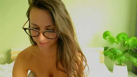 Media: Video of a topless woman with long brown hair, wearing glasses, smiling gently. Background features a green wall with a potted plant and striped cushions.