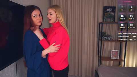 Media: Video of two women in a modern bedroom: one with long, straight red hair in a blue sweater, the other with straight blonde hair in a red sweater, standing close, smiling.