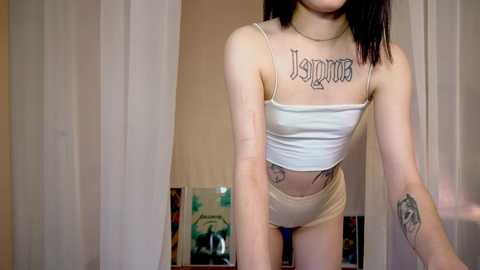 Media: Video of a slim, light-skinned woman with straight black hair, wearing a white crop top and beige shorts, with tattoos on her chest and arms. Background features a white curtain and a cluttered room.