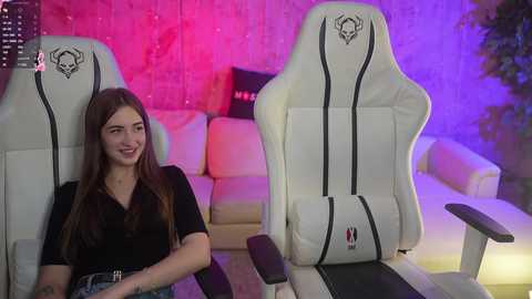 Media: Video of a young woman with long brown hair, smiling, sitting in a white gaming chair with a black skull emblem, in a modern living room with purple and pink lighting.