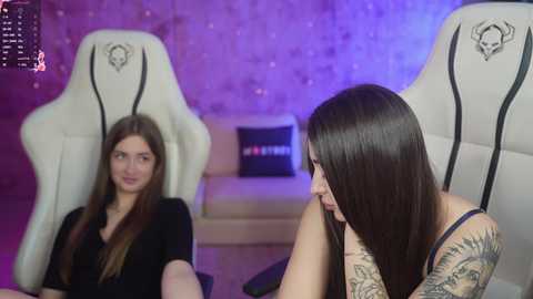 Media: Video of two women in a dimly lit room with purple and blue lighting. One with long brown hair, tattoos, and a black top sits on a white gaming chair. The other, with long brown hair, sits in a similar chair.