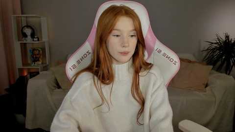 Media: Video of a young woman with long red hair, wearing a white \"SAMSUNG\" gaming chair, sitting in a living room with beige couch, shelves with toys, and potted plants.