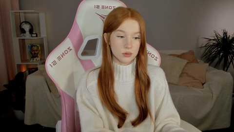 Media: Video of a young, pale-skinned woman with long, straight red hair, wearing a white turtleneck sweater, sitting in a pink and white gaming chair, in a dimly lit living room with beige furniture.