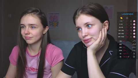 Media: Video of two young girls, one with long brown hair in a pink shirt, the other with short brown hair in a black shirt, sitting in a dimly lit room with posters on the wall.