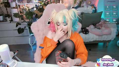 Media: Video of a pale-skinned, blonde-haired woman with pink and blue hair, wearing an orange jacket, black leggings, and a black top, sitting on a white furry chair, holding a phone, in a colorful, cozy room.