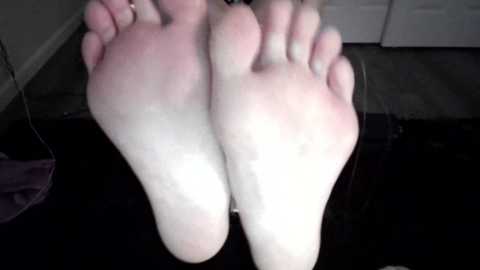 Media: A video of two pale, bare feet with soles facing forward, toes slightly curled, resting on a black surface. The background shows a gray carpet and white door.