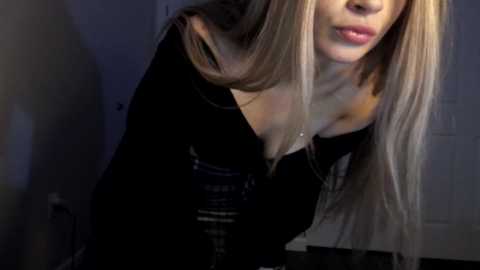 Media: A close-up video of a young Caucasian woman with long, straight blonde hair, wearing a black off-the-shoulder top, leaning forward, lips slightly parted. Background shows a dimly lit room with a door.