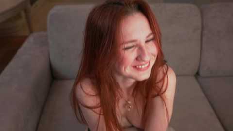 Media: Video of a topless, smiling, fair-skinned woman with long red hair, sitting on a gray couch in a dimly lit room.