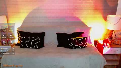 Media: Video of a modern bedroom with a light gray bed featuring black pillows with white patterns, a brick wall backdrop, a glowing lamp, and a red chair.