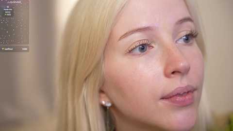 Media: Video of a young, fair-skinned woman with long, straight blonde hair, blue eyes, and subtle makeup. She wears a light-colored dress, and the background is blurred, likely indoors.