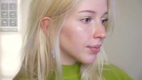 Media: Video of a young Caucasian woman with fair skin, long blonde hair, and blue eyes. She is wearing a green top and is seen in profile against a blurred beige wall.