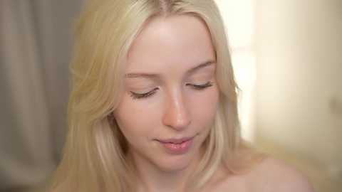 Media: Video of a young Caucasian woman with long, straight, platinum blonde hair, fair skin, and closed eyes, wearing minimal makeup, looking serene. The background is a soft-focus, neutral-toned room with a blurred bed and lamp.