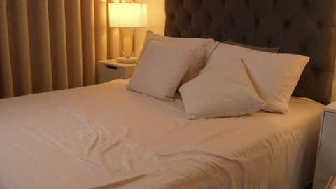 Media: Video of a well-lit, neatly made bed with white sheets, two white pillows, and a dark tufted headboard. A beige curtain covers the left side, and a beige lamp with a white shade sits on a white nightstand.