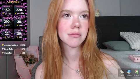 Media: Video of a fair-skinned woman with long, straight red hair, wearing a grey top, looking at the camera in a modern, well-lit bedroom.