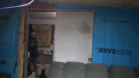 Media: Video of a man in a dark green shirt and khaki shorts standing in a dimly lit, cluttered room with blue tarp walls and a closed door.