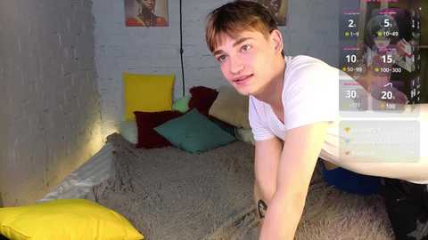 Media: Video of a young, fair-skinned, short-haired man with a slim build, wearing a white T-shirt, leaning forward on a gray bed with colorful pillows in a dimly lit room with white brick walls.