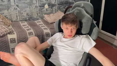 Media: A video of a young boy with light skin and short brown hair, wearing a white t-shirt and shorts, sitting in a gaming chair in a bedroom with patterned wallpaper and a bed.