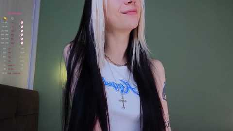 Media: Video of a young woman with long, straight black hair with white streaks, wearing a white tank top with a blue tattoo, standing against a green wall.