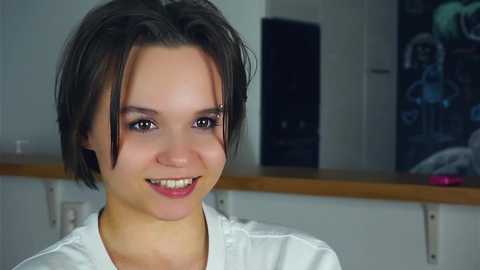 Media: Video of a young woman with short, dark brown hair, fair skin, and a white top, smiling. Background features a modern kitchen with white cabinets and a dark countertop.