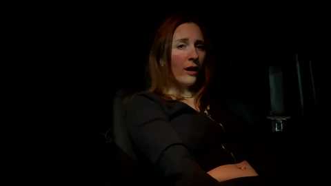 Media: A dimly lit video of a woman with shoulder-length auburn hair, wearing a black blouse, and a chain necklace, seated in a dark room, with a lit candle and wine glass in the background.