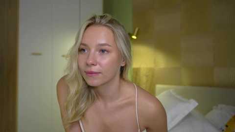 Media: Video of a blonde woman with fair skin and a slim physique, wearing a white camisole, sitting on a bed in a softly lit bedroom.