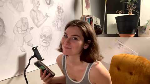 Media: Video of a young woman with shoulder-length brown hair, wearing a white tank top, sitting in a mustard-yellow chair. She holds a black microphone, surrounded by sketchbook drawings on a white wall, with a potted plant on the shelf behind her.