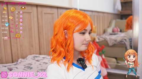 Media: Video of a young woman with orange wavy hair, wearing a white and blue striped shirt, standing in a kitchen with wooden cabinets. The background features a cluttered countertop and a chibi anime character.