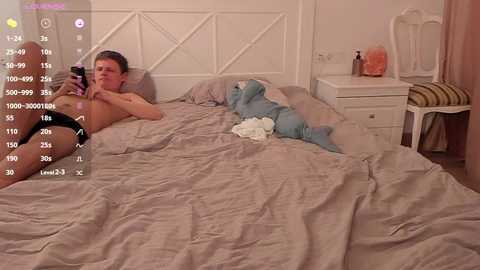 Media: Video of a young man in black underwear lying on a bed, taking a selfie. The room features a white headboard, a nightstand with a lamp, and a white chair.