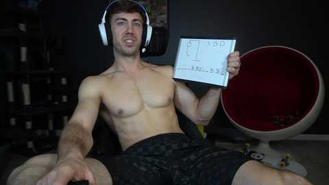 Media: Video of a shirtless, muscular, white man with short brown hair, wearing headphones and holding a white paper with numbers, seated in a dark room with a red egg chair and shelves in the background.