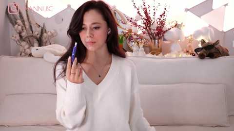 Media: Video of a young woman with long dark hair, fair skin, and a slender physique, wearing a white V-neck sweater, holding a blue pen, standing in a softly lit, minimalist room with white furniture and decorative items.