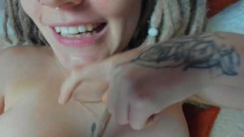 Media: Video of a smiling, topless woman with light skin, blonde dreadlocks, and a large tattoo on her arm. She gestures with her fingers. Background is a blurred red and white setting.