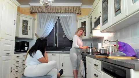 Media: Video of a modern kitchen with two women: one in white leggings and a pink top, the other in a gray dress, cooking. White cabinets, dark countertop, purple bag, and a window with sheer curtains.