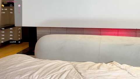 Media: A video of a minimalist bedroom with a beige leather headboard and a wooden nightstand on the left. The background is a plain white wall with a red accent strip. A bed with cream-colored sheets is partially visible.