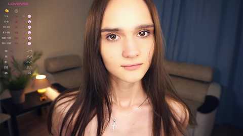 Media: Video of a young woman with long brown hair, fair skin, and a slender physique, wearing a necklace, in a cozy living room with beige furniture and a lit table lamp.