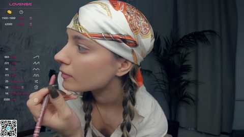 Media: Video of a young woman with light skin, braided hair, wearing a white shirt and orange headscarf, applying makeup. Background shows a calendar and a plant.