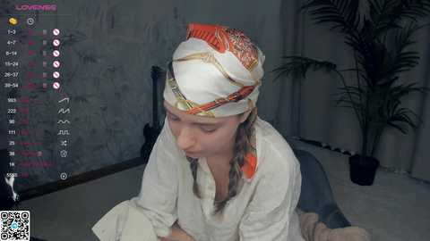 Media: Video of a young woman with fair skin and long brown hair, wearing a white robe, sitting on a chair in a dimly lit room. She has a colorful headscarf with red, orange, and green patterns.