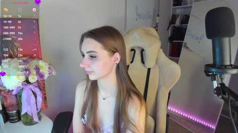 Media: Video of a young woman with long brown hair, wearing a sleeveless top, sitting in a beige gaming chair. The room features a flower vase, calendar, and a microphone stand.