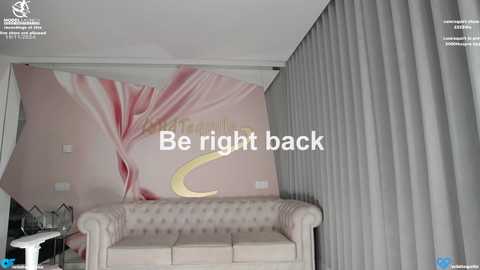 Media: Video of a modern living room with a beige tufted sofa, gray curtains, and a large, vibrant pink and white floral mural on the wall. Text overlay reads \"Be right back.\