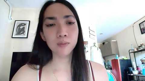 Media: Video of a young Asian woman with long black hair and fair skin, wearing a red tank top, standing in a bright kitchen with white walls and framed artwork.