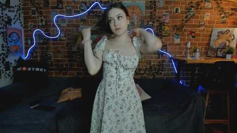 Media: Video of a young woman with shoulder-length brown hair, wearing a floral dress, standing in a dimly lit room with a brick wall, neon blue light, and a black couch.