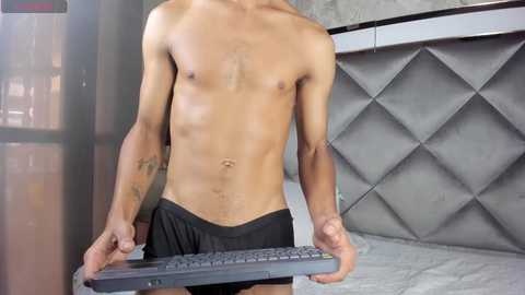 Media: Video of a shirtless, fit male with light skin and tattoos, wearing black briefs, holding a black remote control, standing in front of a gray, diamond-patterned wall in a modern room.