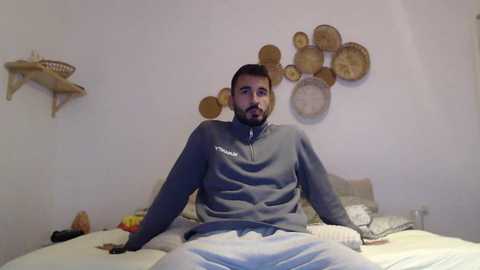 Media: A video of a bearded man with short dark hair wearing a gray sweatshirt and white pants sitting on a bed with white sheets, surrounded by wooden wall decorations and a cat-shaped shelf in a minimalist, light-colored room.