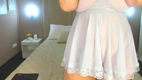 Media: A video of a woman in a sheer white nightgown, standing in a modern bedroom with beige walls and a bed, featuring a white pillow and a nightstand with a vase of yellow flowers.