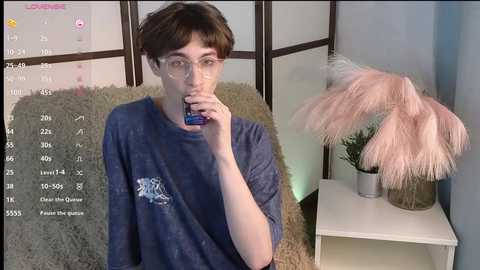 Media: Video of a young, slender person with short brown hair, wearing glasses, a blue t-shirt, and drinking from a blue bottle, in a room with a calendar, fluffy rug, pink feather duster, and a white side table.