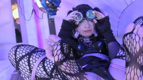 Media: Video of a pale-skinned, androgynous individual with black hair, wearing fishnet tights, black leather jacket, and mirrored goggles, reclining provocatively on a white leather couch, surrounded by purple and blue lighting.