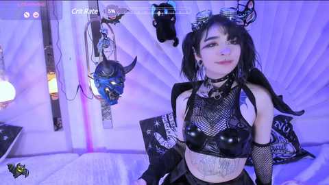 Media: Video of a young woman with pale skin, black hair in pigtails, wearing a black leather crop top, fishnet sleeves, and a choker. She stands in a room adorned with white drapes, a skull mask, and a glowing lamp.