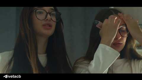 Media: Video of two young women, one with glasses, adjusting the other's glasses. Both have long hair, fair skin, and wear white tops. Background is blurred, focusing on their faces.