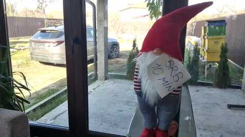 Media: A video of a large, whimsical elf figurine with a red hat and white beard, holding a sign that reads \"Merry Christmas,\" positioned on a glass window sill overlooking a suburban street with parked cars and a yellow dumpster.