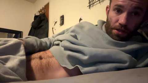 Media: Video of a bearded man lying on a bed in a dimly lit room, wearing a grey sweater, exposing his bare stomach.