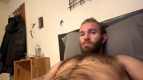 Media: Video of a shirtless, bearded Caucasian man with light skin and a muscular build, reclining on a gray couch. Background includes a wooden side table, a water bottle, and a hanging jacket.